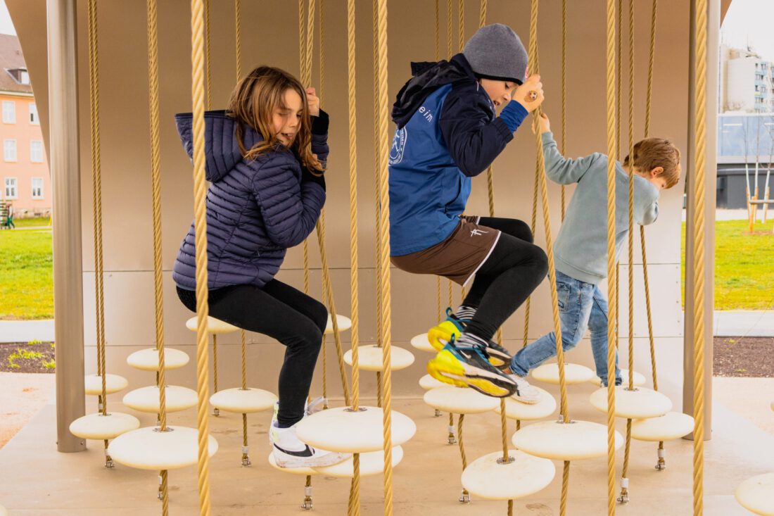 Spiel-und-Bewegungspark-auf-Spinelli-Gelände der ©-BUGA-23, fotografiert von Carina-Drexler. Drei Kinder bewegen sich zwischen schwankenden, runden "Tellern", die in verschiedener Höhe an senkrechten Seilen montiert sind und haben sichbar Spaß.